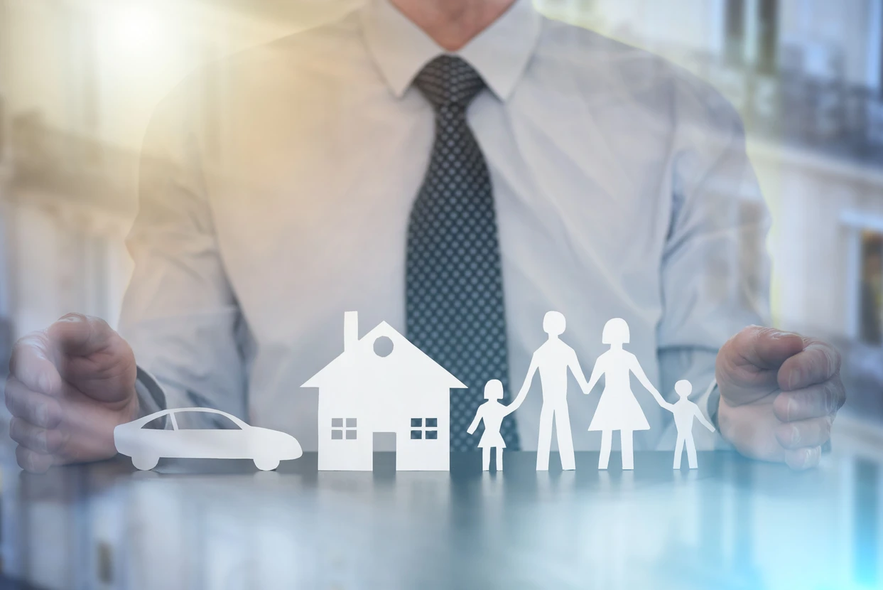 Homme d'affaires en chemise blanche et cravette protégeant de ses mains des silhouettes blanches représentant une voiture, une maison et une famille avec deux enfants, symbolisant les services d'assurance et de protection