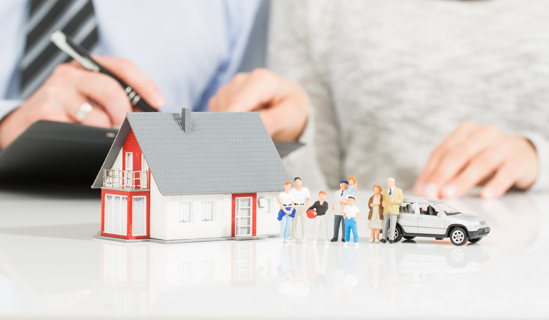 Personne en chemise bleue protégeant de ses mains une famille en papier découpé blanc composée de quatre silhouettes et une maison miniature avec fenêtres, symboles d'assurance familiale et protection du foyer
